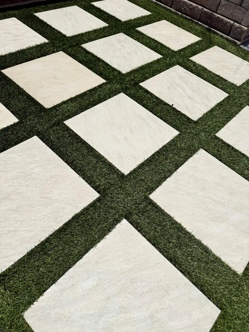 Close-up of artificial turf strips between square porcelain pavers set on the diagonal, with a block retaining wall behind
