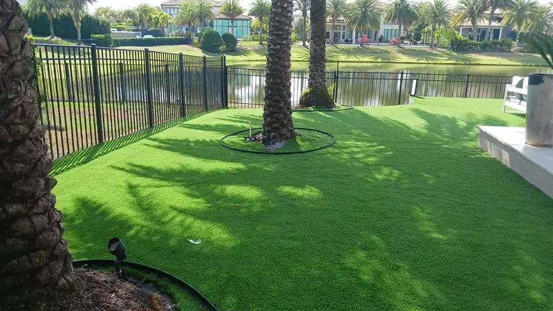 Lakefront backyard with artificial turf, two palms inside black ring edging, an aluminum picket fence and homes across the water