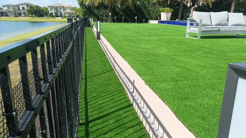 Large lakefront yard covered in artificial turf, running from an aluminum picket fence past path lights to a pool, an outdoor sofa and putting green flags