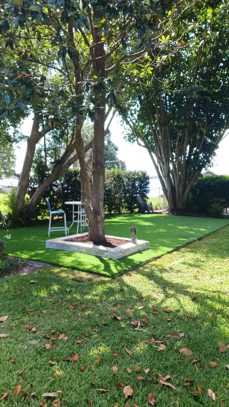 Small artificial turf seating area with a bistro table around a raised stone planter under a magnolia, natural grass in front and a lake behind